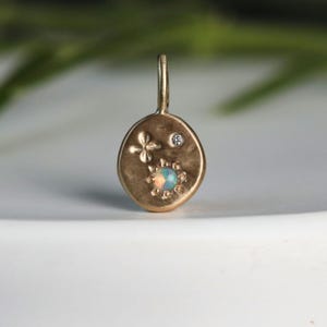 Puede incluir: Colgante dorado con un lazo. Presenta una flor de cuatro pétalos, un pequeño diamante y una piedra de ópalo rodeada de cuentas doradas. El colgante tiene una superficie texturizada. El ópalo es azul y blanco.
