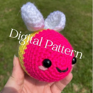 Puede incluir: Una abeja de ganchillo hecha a mano con un cuerpo rosa brillante, detalles amarillos y alas blancas. La abeja tiene ojos de cuentas negras y una sonrisa cosida. El texto "Digital Pattern" está superpuesto.