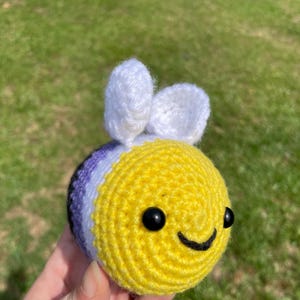 May include: A crocheted bee toy with a yellow body, black eyes, and a smiling mouth. The bee has white antennae and a section in the non-binary pride flag colours: purple, white, and black.