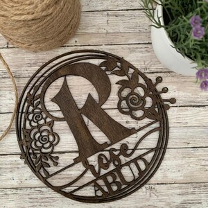 Puede incluir: Un círculo de madera marrón con un recorte de la letra R y diseños florales. El texto "Rose" y "Abud" también están recortados en la madera.