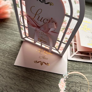 May include: A pink and white paper cut-out card with a floral design. The card is folded in half and has a window cut-out in the front. The text "Luci's Quince Años" is written in gold lettering on the front of the card.