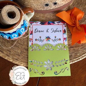 May include: A green card with a white border and a cut-out design. The card has the text "Diane & Joshua" in a colorful floral pattern. The card is on a brown woven surface with a straw hat and a bright orange bow.