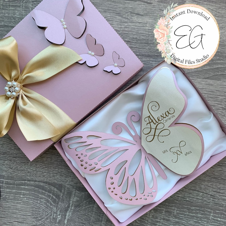 May include: A pink gift box with a gold ribbon and butterfly decorations. The box is open to reveal a white satin-lined box with a pink butterfly cutout and a white card with the text "Alexa Carranza Mis 7 A&ntilde;os".