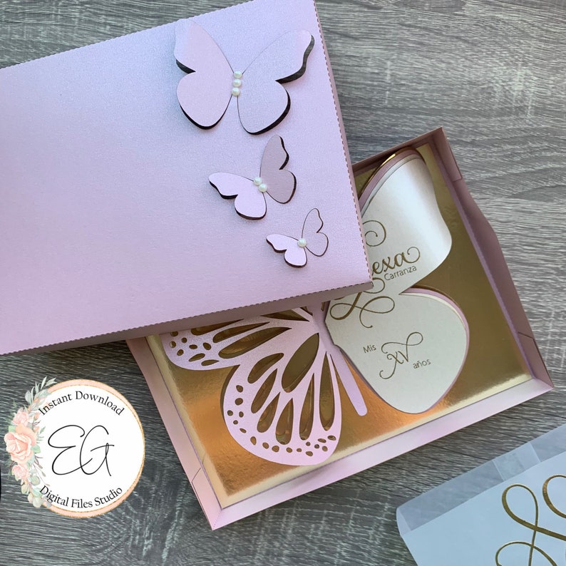 May include: A pink and gold butterfly-themed box with a white card inside. The card has the text "Alexa Carranza" and "Mis XV a&ntilde;os".