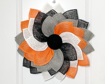 May include: A black, orange, white, and gray burlap wreath in the shape of a flower. The wreath is hanging on a white door.