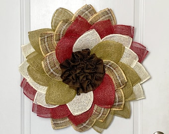 May include: A decorative sunflower wreath made of burlap in shades of red, brown, cream, and green. The wreath is hanging on a white door. The center of the sunflower is a dark brown, ruffled material.