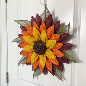 May include: A burlap sunflower wreath with layers of burgundy, orange, and yellow petals. The center of the sunflower is brown and the leaves are green.