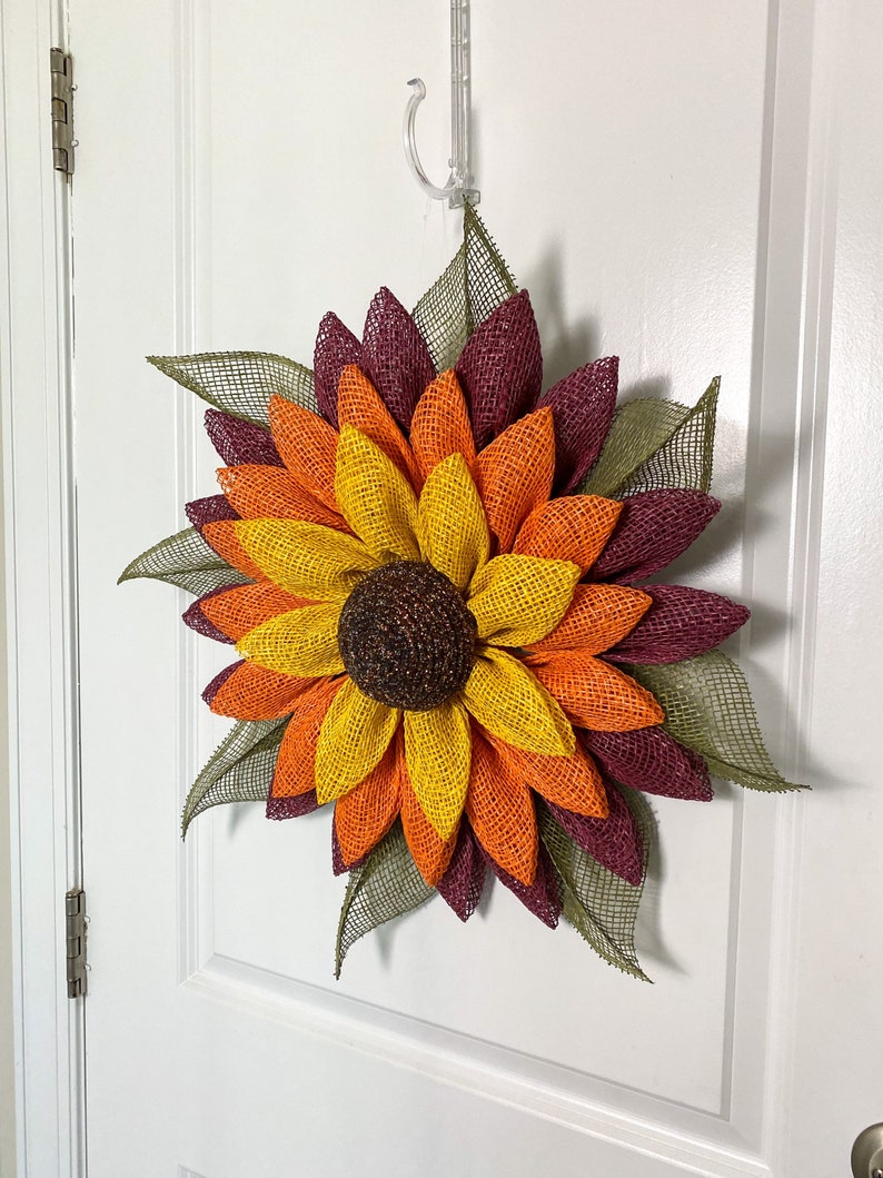 May include: A burlap sunflower wreath with layers of burgundy, orange, and yellow petals. The center of the sunflower is brown and the leaves are green.