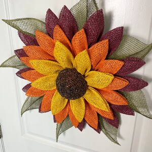 May include: A burlap sunflower wreath with layers of burgundy, orange, and yellow petals. The center of the sunflower is brown with glitter accents. The wreath is decorated with green burlap leaves.