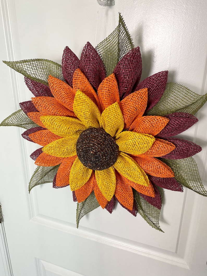 May include: A burlap sunflower wreath with layers of burgundy, orange, and yellow petals. The center of the sunflower is brown with glitter accents. The wreath is decorated with green burlap leaves.