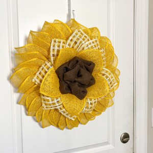 May include: A yellow and brown sunflower wreath made of burlap mesh. The wreath has a brown burlap center and white and yellow checkered accents.