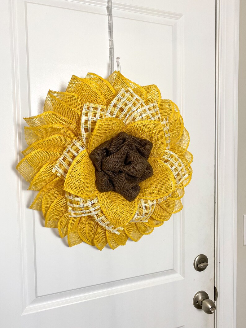 May include: A yellow and brown sunflower wreath made of burlap mesh. The wreath has a brown burlap center and white and yellow checkered accents.