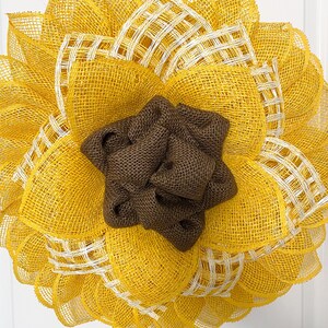 May include: A yellow and brown sunflower wreath made of burlap and mesh. The wreath has a brown burlap center and yellow mesh petals with white accents.