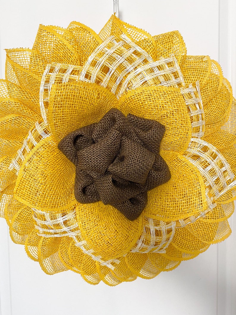 May include: A yellow and brown sunflower wreath made of burlap and mesh. The wreath has a brown burlap center and yellow mesh petals with white accents.