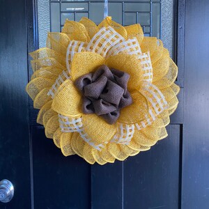 May include: A yellow and white burlap wreath with a brown burlap center, resembling a sunflower. The wreath is hanging on a black door.