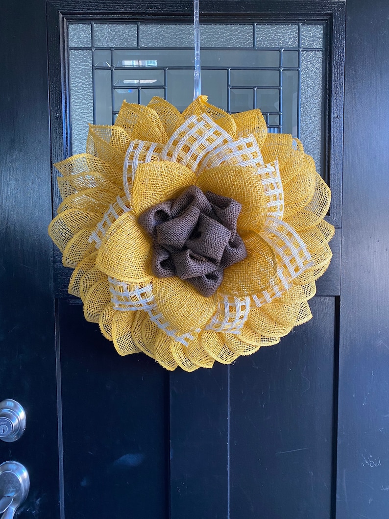 May include: A yellow and white burlap wreath with a brown burlap center, resembling a sunflower. The wreath is hanging on a black door.