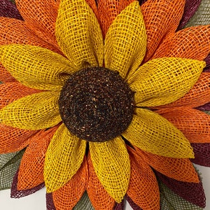 May include: A large sunflower wreath made of burlap mesh in shades of burgundy, orange, and yellow. The center of the wreath is a brown and gold glitter circle.