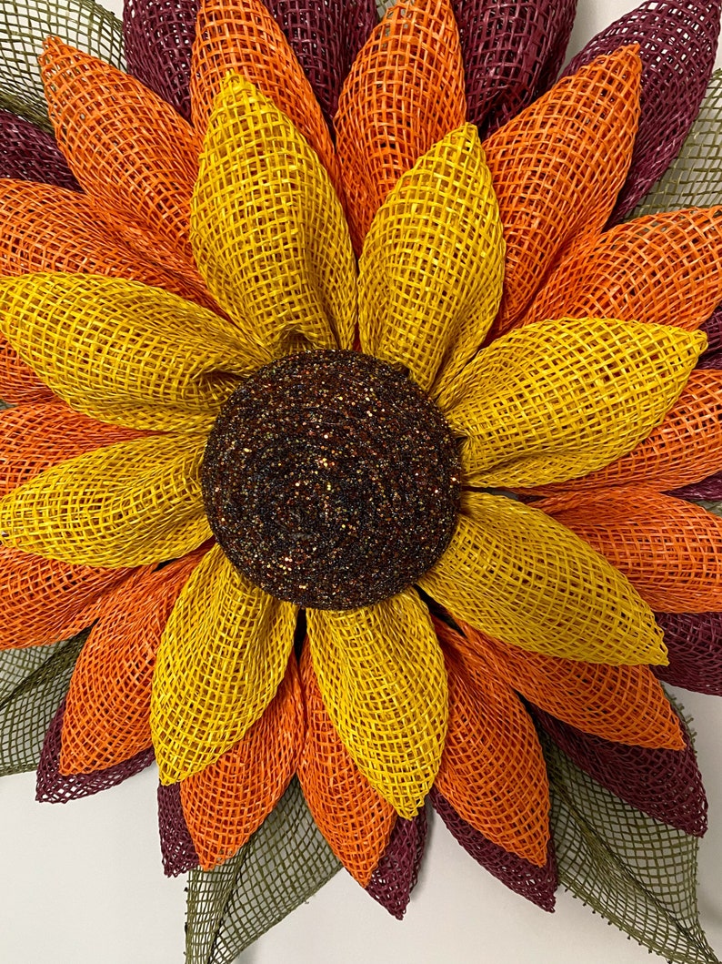 May include: A large sunflower wreath made of burlap mesh in shades of burgundy, orange, and yellow. The center of the wreath is a brown and gold glitter circle.