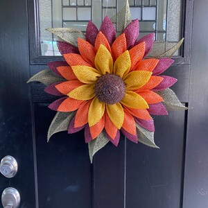 May include: A fall-themed sunflower wreath with burgundy, orange, and yellow burlap petals. The wreath is made of burlap and has a brown center.