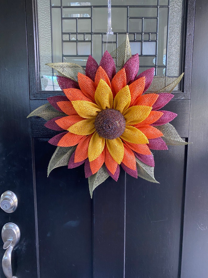 May include: A fall-themed sunflower wreath with burgundy, orange, and yellow burlap petals. The wreath is made of burlap and has a brown center.