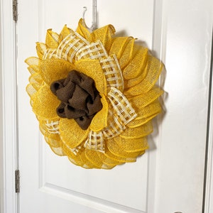 May include: A yellow and white sunflower wreath with a brown burlap center. The wreath is hanging on a white door.