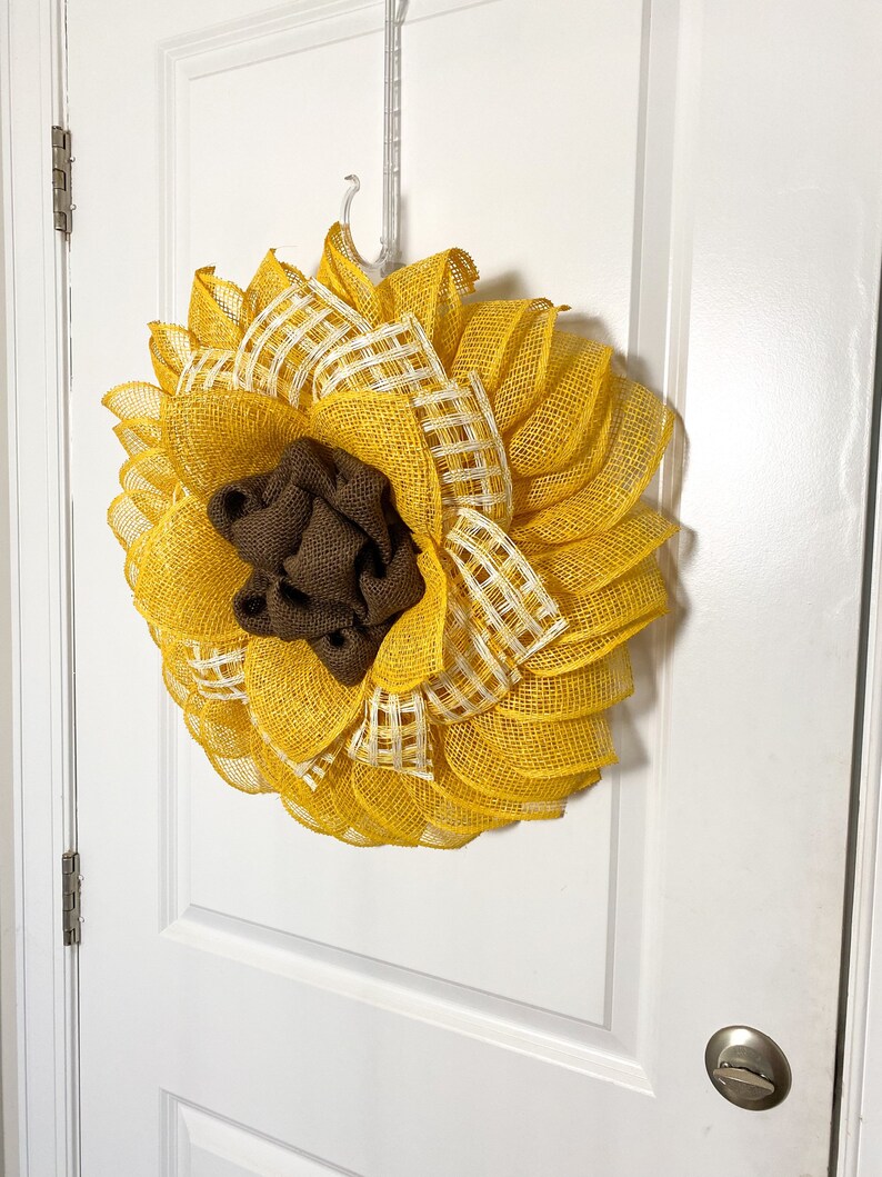 May include: A yellow and white sunflower wreath with a brown burlap center. The wreath is hanging on a white door.