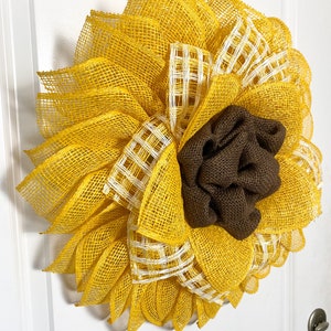 May include: A yellow and brown sunflower wreath made of burlap and mesh. The wreath has a brown burlap center and yellow mesh petals with white checkered accents.