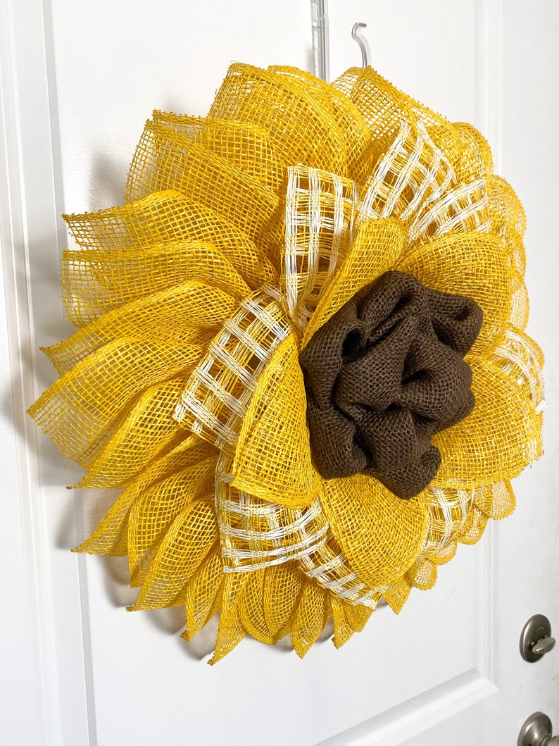 May include: A yellow and brown sunflower wreath made of burlap and mesh. The wreath has a brown burlap center and yellow mesh petals with white checkered accents.