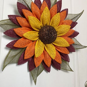 May include: A large, handmade sunflower wreath with burgundy, orange, and yellow burlap petals. The center of the sunflower is brown and has a glittery texture.