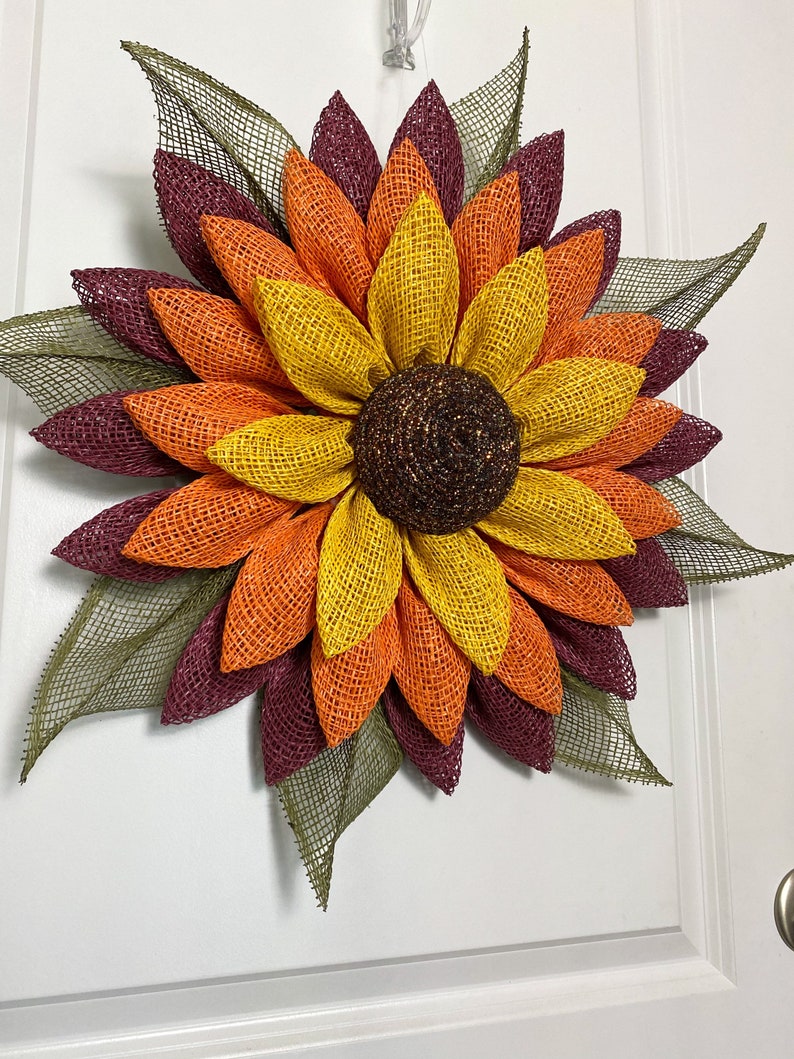 May include: A large, handmade sunflower wreath with burgundy, orange, and yellow burlap petals. The center of the sunflower is brown and has a glittery texture.