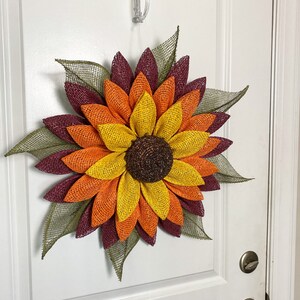 May include: A large sunflower wreath with burgundy, orange, and yellow burlap petals. The wreath is hanging on a white door.