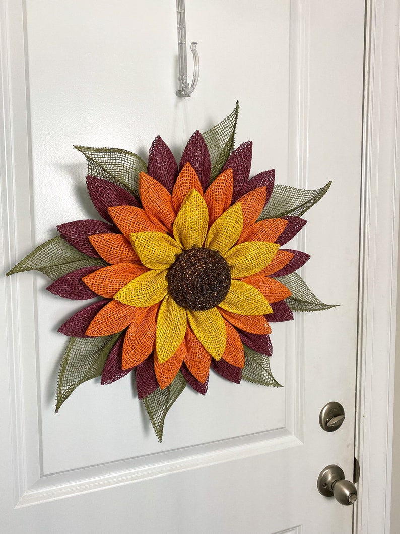 May include: A large sunflower wreath with burgundy, orange, and yellow burlap petals. The wreath is hanging on a white door.