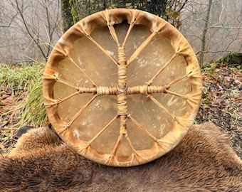 May include: A round, handcrafted drum with a natural hide drumhead and a woven handle. The drum is tan and brown, with a circular frame and a cross-shaped handle. The drum is resting on a brown, textured fur.