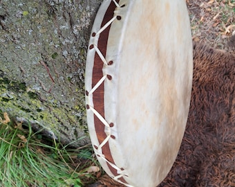 May include: A round, hand-held drum with a natural hide head and a dark brown wooden frame. The drum is laced with white cord and rests against a tree trunk and a brown fur rug. The drum's surface has a light tan color.