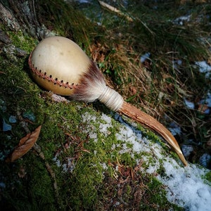 May include: A handcrafted maraca with a light tan gourd body, stitched with red thread. The handle is made of wood and the base of the gourd is covered with brown fur. The maraca rests on a bed of green moss and snow.