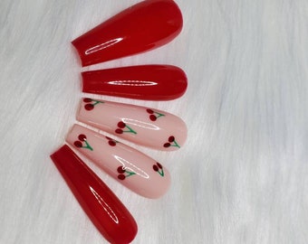 May include: A set of five artificial nails. Two are solid red, and three are light pink with cherry designs. The nails are rectangular with rounded tips, and are arranged on a white, fluffy surface.