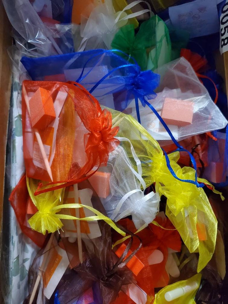 May include: A collection of small, colorful organza bags filled with various nail care items. The bags are in shades of red, orange, yellow, blue, green, white, and brown. The contents include orange nail buffers and wooden sticks.