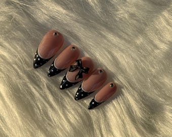 May include: Almond-shaped artificial nails with a nude base, black tips with white polka dots, and a black bow accent. Each nail has a small, gold-colored stud. The nails are arranged on a white, fluffy surface.