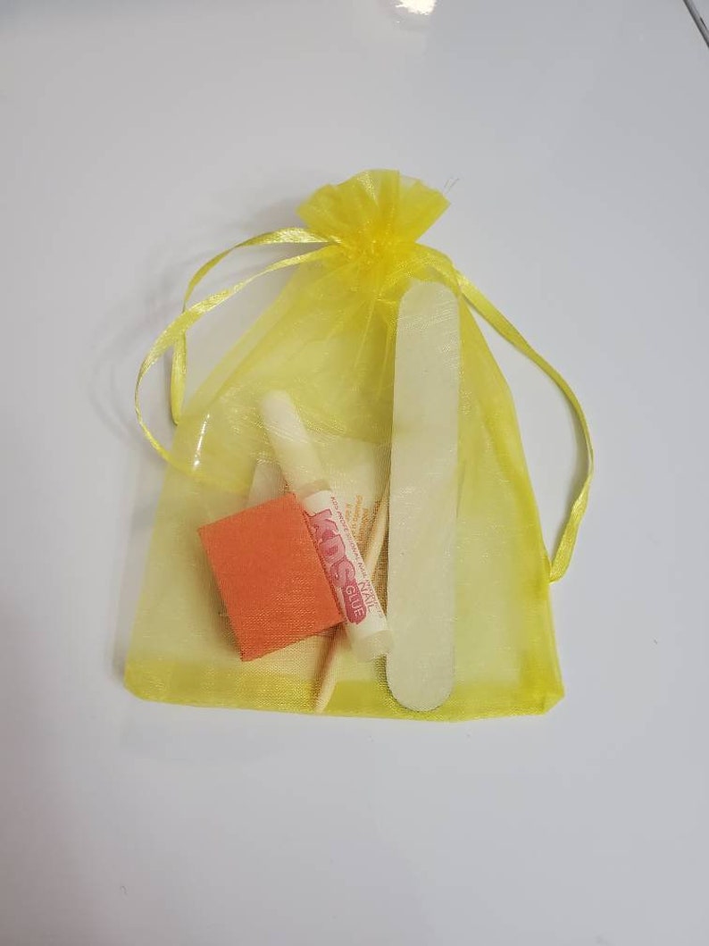 May include: A yellow organza drawstring bag containing nail care tools. The bag holds a nail file, a small orange buffer, a wooden cuticle stick, and a tube of nail glue. The nail glue tube has the text "KDS NAIL GLUE".