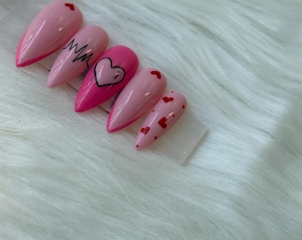 May include: Set of five pink press-on nails with a glossy finish. Designs include a small red heart, a black squiggly line, a black heart outline, and red heart accents. The nails are displayed on a white, fluffy surface.