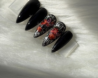 May include: A set of black, almond-shaped press-on nails. Two nails feature a floral design with red and white flowers on a black and white grid pattern. The other nails are solid black.