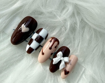 May include: A set of five artificial nails with various designs. One nail features a white heart on a dark brown base. Another has a black and white checkerboard pattern. Other nails have chocolate drizzle, a white bow, and a small heart.