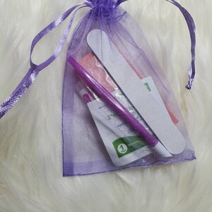 May include: A purple organza gift bag containing nail care tools. The bag holds a nail file, a purple cuticle pusher, and other manicure supplies. The bag is tied with a matching purple ribbon, and the contents are visible through the sheer fabric.
