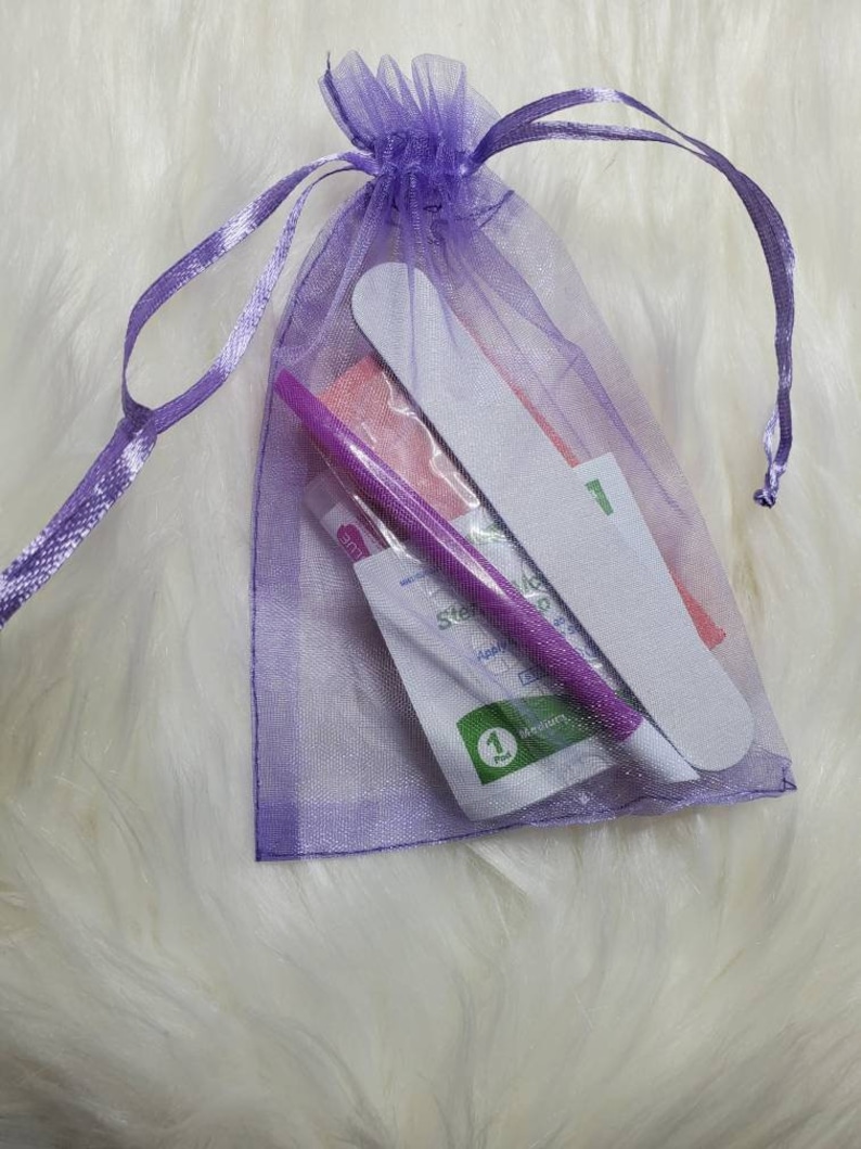 May include: A purple organza gift bag containing nail care tools. The bag holds a nail file, a purple cuticle pusher, and other manicure supplies. The bag is tied with a matching purple ribbon, and the contents are visible through the sheer fabric.
