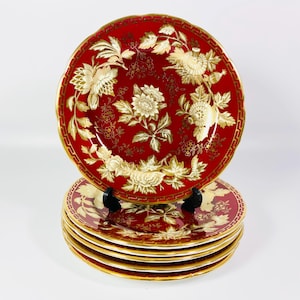 May include: A set of six red and gold decorative plates. Each plate features a floral design with gold accents and a gold rim. The plates are stacked on a white surface.