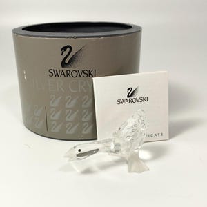 Può includere: Una statuetta di cigno in cristallo trasparente con becco e occhio neri, appoggiata su una base trasparente. La statuetta è accanto a una scatola cilindrica grigia e a una carta bianca, entrambe con il logo Swarovski.