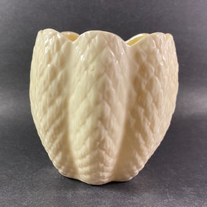 May include: A cream-coloured ceramic vase with a textured, quilted pattern. The vase has a scalloped rim and a rounded shape. The neutral colour and unique texture make it a versatile decorative piece for any home.