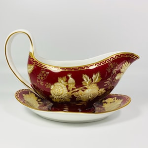 Op de afbeelding: Een rood en goudkleurig jusvat met bijpassende schotel. Het jusvat heeft een gebogen handvat en een decoratief gouden bloemmotief. De schotel is rond en heeft ook een gouden bloemdesign. De rand van het jusvat en de schotel is goudkleurig.