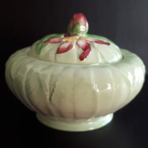 May include: A green ceramic sugar bowl with a lid featuring a red and yellow flower design. The bowl has a ribbed texture and a rounded shape.