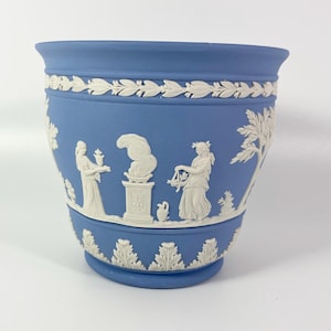 Può includere: Un vaso per piante in ceramica blu opaco con decorazioni in rilievo bianche. Il design presenta un bordo superiore a foglie di alloro, una fascia inferiore a foglie d'acanto e un fregio centrale con due figure vicino a un altare fumante.